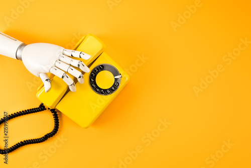 cropped shot of robotic hand reaching for vintage phone on yellow tabletop