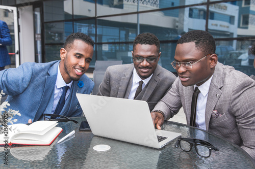 Group of happy diverse male business people afro american team in formal gathered around laptop computer in bright office against the background of a glass building