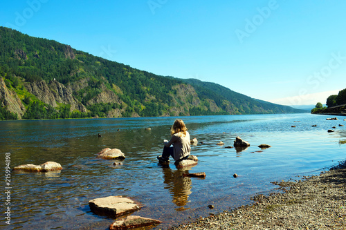 The blonde girl sits on a rocky shore and looks into the distance to the sea and high mountains.