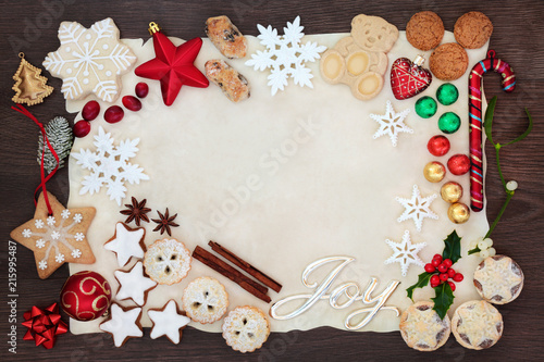 Christmas background border with joy sign including tree decorations, biscuits, cakes, chocolates and winter flora on parchment paper on rustic oak. Flat lay.