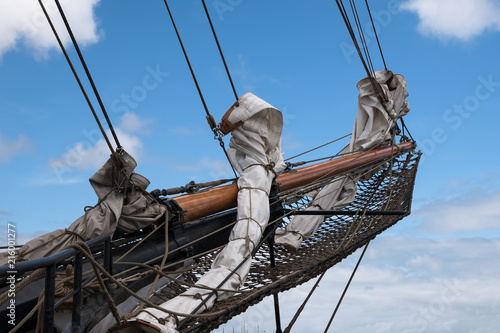 bowsprit and jib boom with reefed sails on the bow of a historic sailing ship against a blue sky with clouds