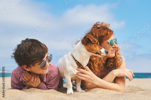Happy mother with her son and their dog breed Jack Russell lie on the sand on a large beach by the sea and look at the sun in the summer. Family vacation on summer vacation. Home pet