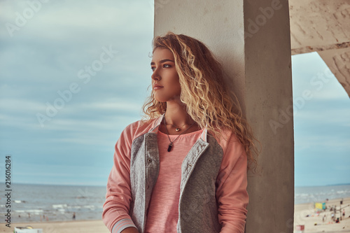 Portrait of a smiling charming young girl with blonde hair dressed in a pink jacket leaning on a concrete column against sea coast.