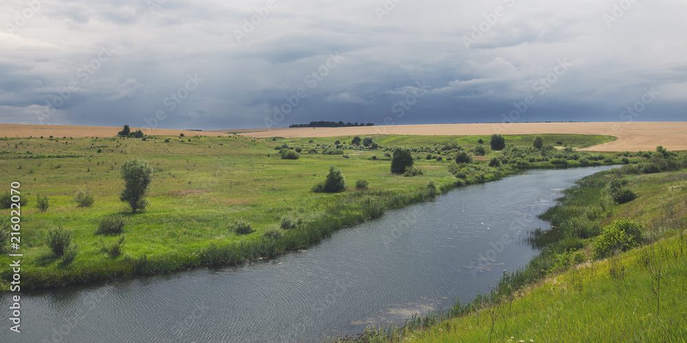 Cloudy summer landscape with wheat fields,meadows,woods and small river.Dark stormy clouds in dramatic overcast sky.