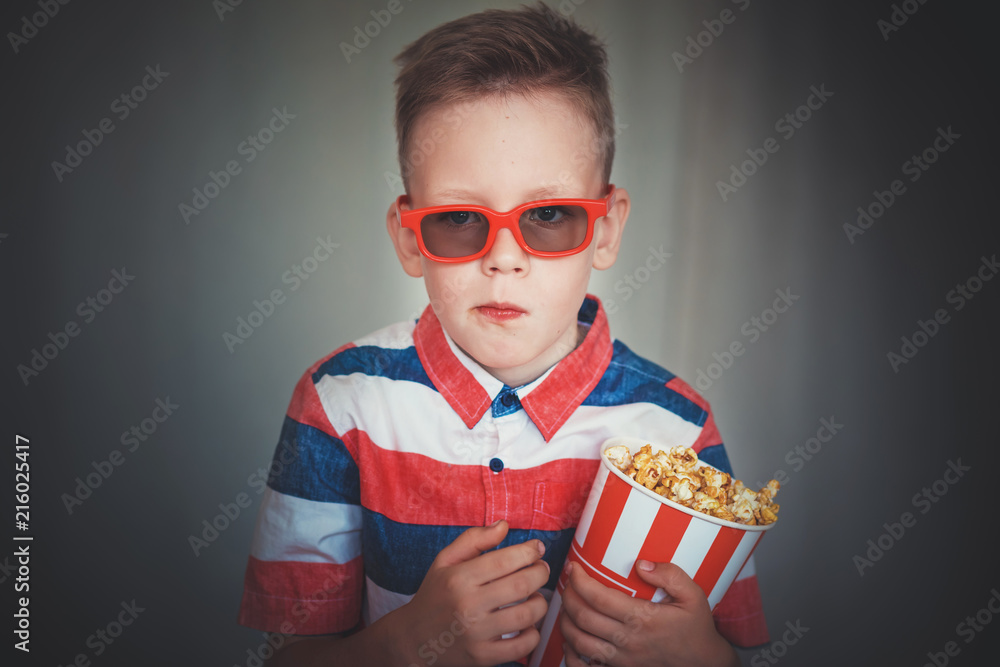 Fototapeta premium Young boy watch a movie in 3D glasses at the cinema or at home. Little kid eat popcorn over gray background. Home theater. Cute Child in vintage cinema eyeglasses