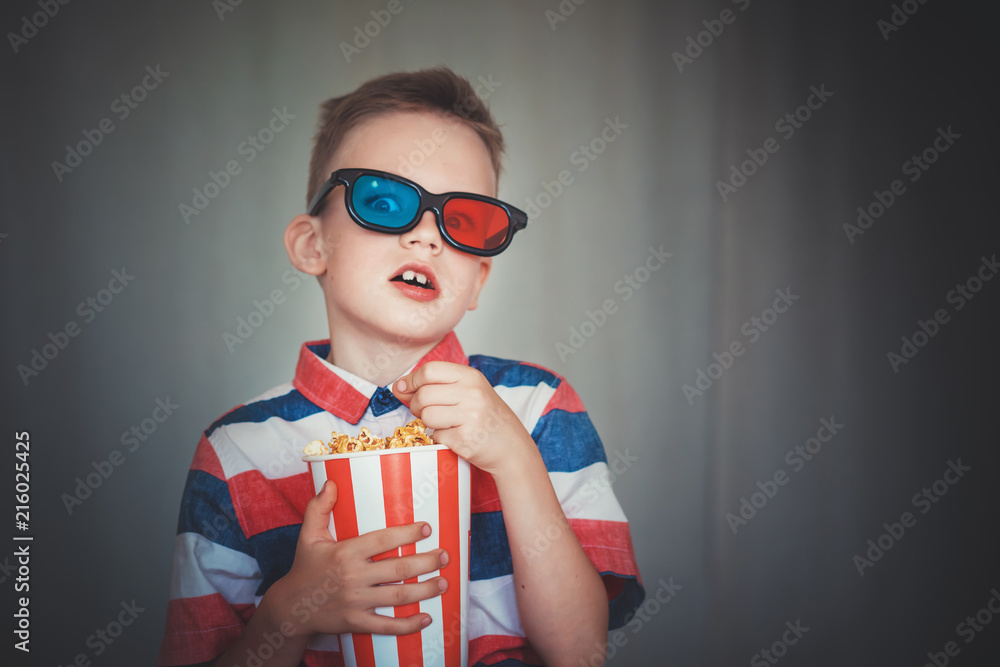 Young boy watch a movie in 3D glasses at the cinema or at home. Little kid  eat popcorn over gray background. Home theater. Cute Child in vintage  cinema eyeglasses Stock Photo