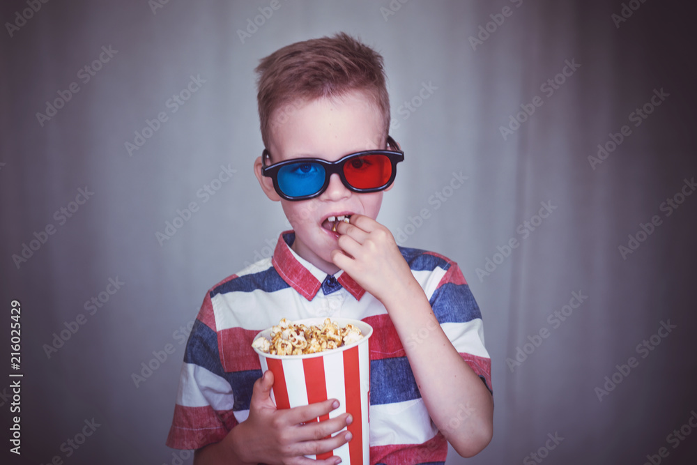 Fototapeta premium Young boy watch a movie in 3D glasses at the cinema or at home. Little kid eat popcorn over gray background. Home theater. Cute Child in vintage cinema eyeglasses
