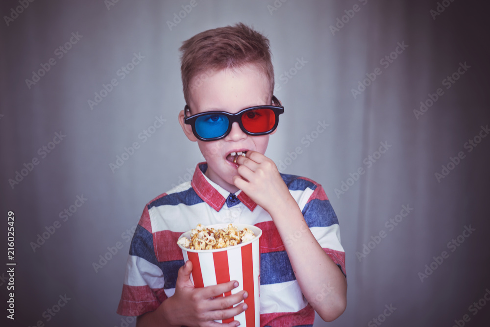 Young boy watch a movie in 3D glasses at the cinema or at home. Little kid eat popcorn over gray background. Home theater. Cute Child in vintage cinema eyeglasses