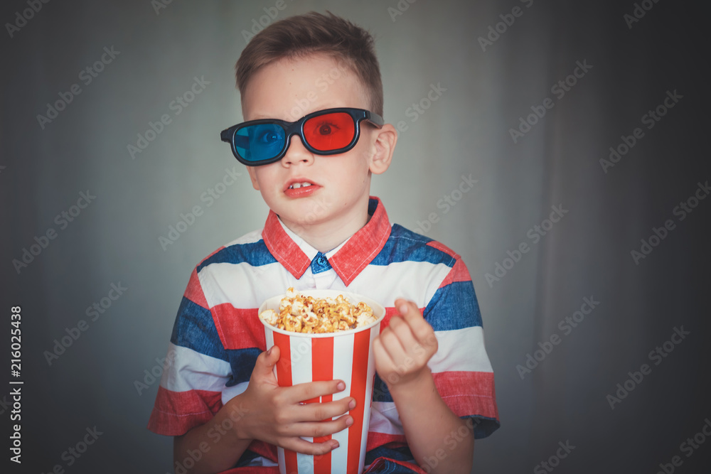 Fototapeta premium Young boy watch a movie in 3D glasses at the cinema or at home. Little kid eat popcorn over gray background. Home theater. Cute Child in vintage cinema eyeglasses