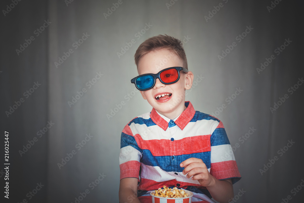 Fototapeta premium Young boy watch a movie in 3D glasses at the cinema or at home. Little kid eat popcorn over gray background. Home theater. Cute Child in vintage cinema eyeglasses