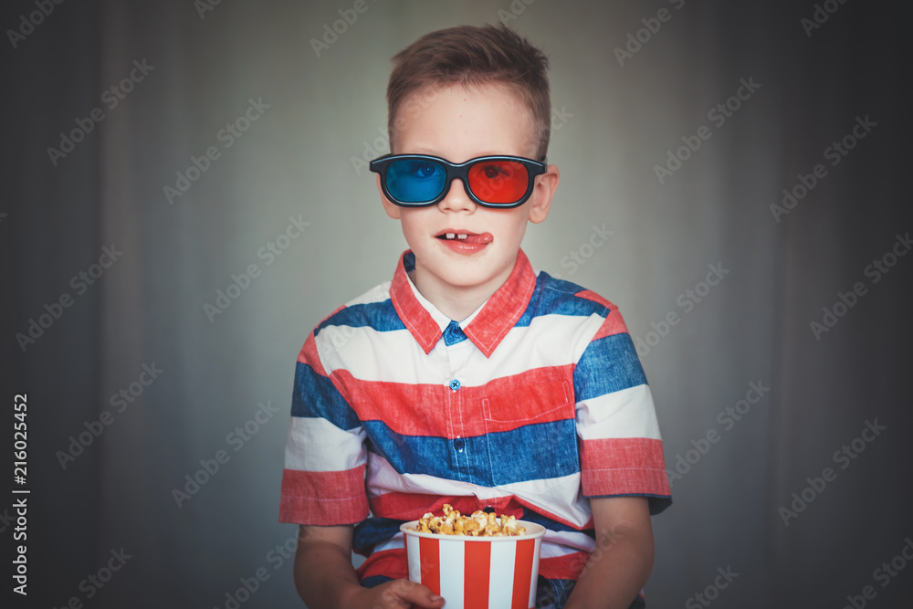 Fototapeta premium Young boy watch a movie in 3D glasses at the cinema or at home. Little kid eat popcorn over gray background. Home theater. Cute Child in vintage cinema eyeglasses