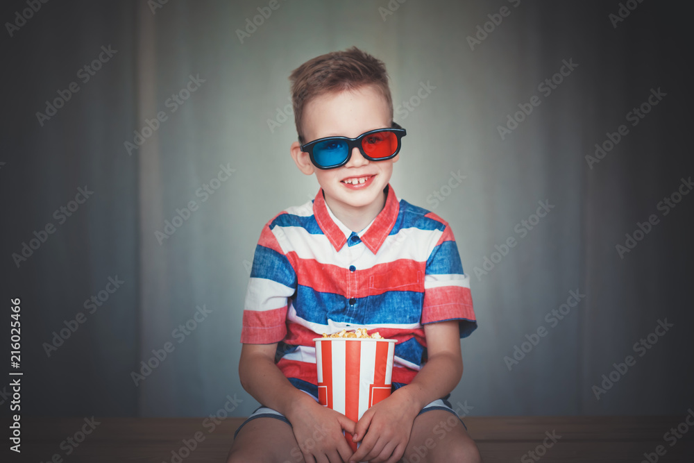 Fototapeta premium Young boy watch a movie in 3D glasses at the cinema or at home. Little kid eat popcorn over gray background. Home theater. Cute Child in vintage cinema eyeglasses