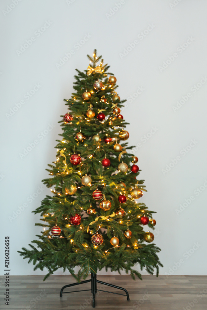 Beautiful Christmas tree with fairy lights and festive decor near white wall Beautiful Christmas tree with fairy lights and festive decor near white wall