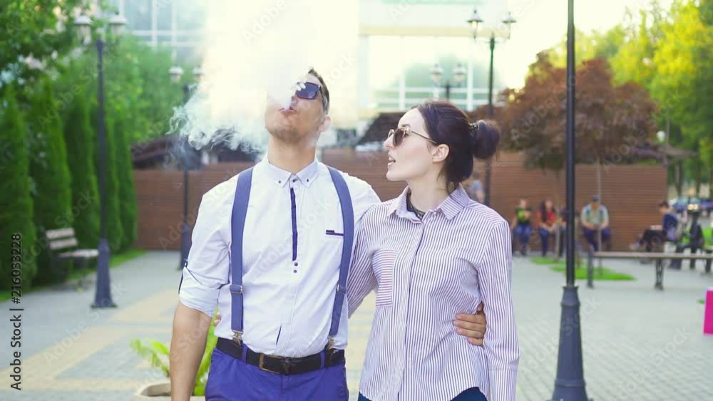 Stylish man with e cigarette and girl walking in the park,sunflare.Slow mo Stylish man with e cigarette and girl walking in the park,sunflare.Slow mo