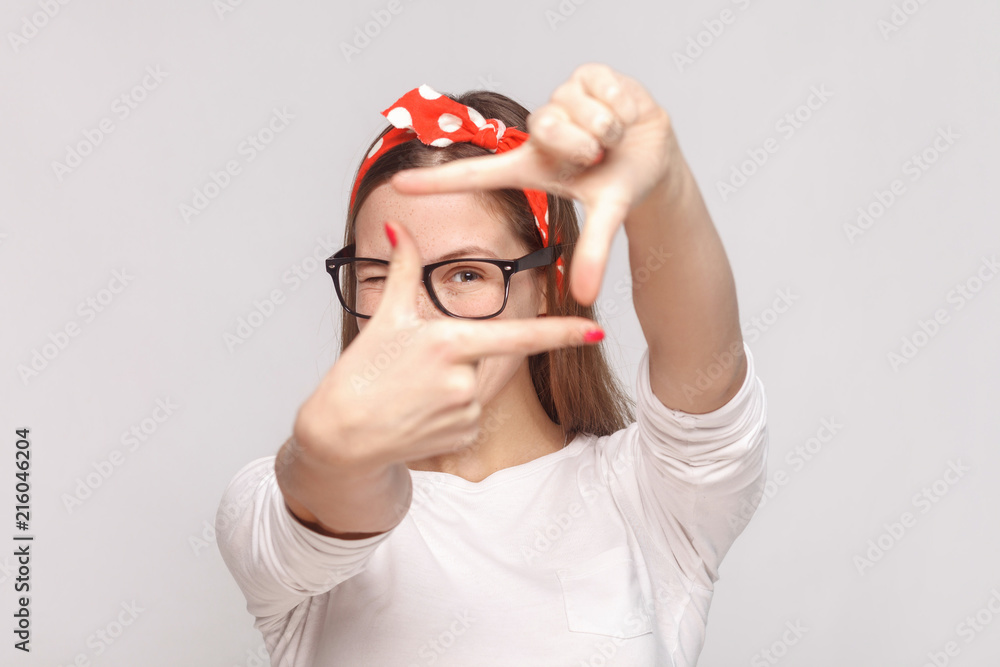 Fototapeta premium choosing composition or focusing portrait of beautiful emotional young woman in white t-shirt with freckles, glasses, red lips and head band. indoor studio shot, isolated on light gray background.