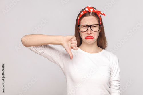 Fotografie, Obraz its bad, sad thumbs down of unsatisfied emotional young woman in white t-shirt with freckles, black glasses, red lips and head band looking at camera