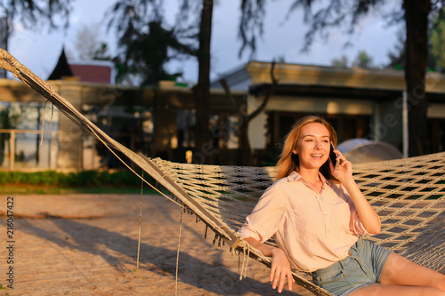 Young european woman sitting in hammock and talking by smartphone, summer vacations. Concept of resting in tropicak resort and advantageous tariff plan, modern technology. photo