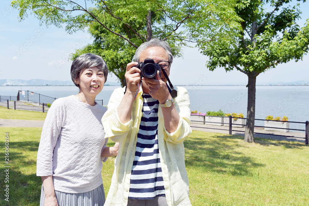 Φωτογραφία 海の近くの公園で、笑顔でカメラを構えるシニア夫婦
