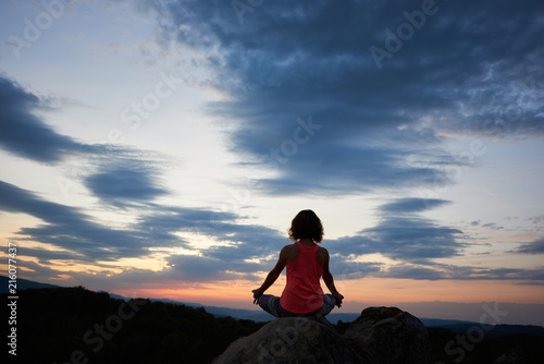 Back view of young slim woman sitting on big rock in yoga lotus pose on green tree tops dramatic evening sky at sunset background. Tourism, meditation and healthy lifestyle concept. Siddhasana