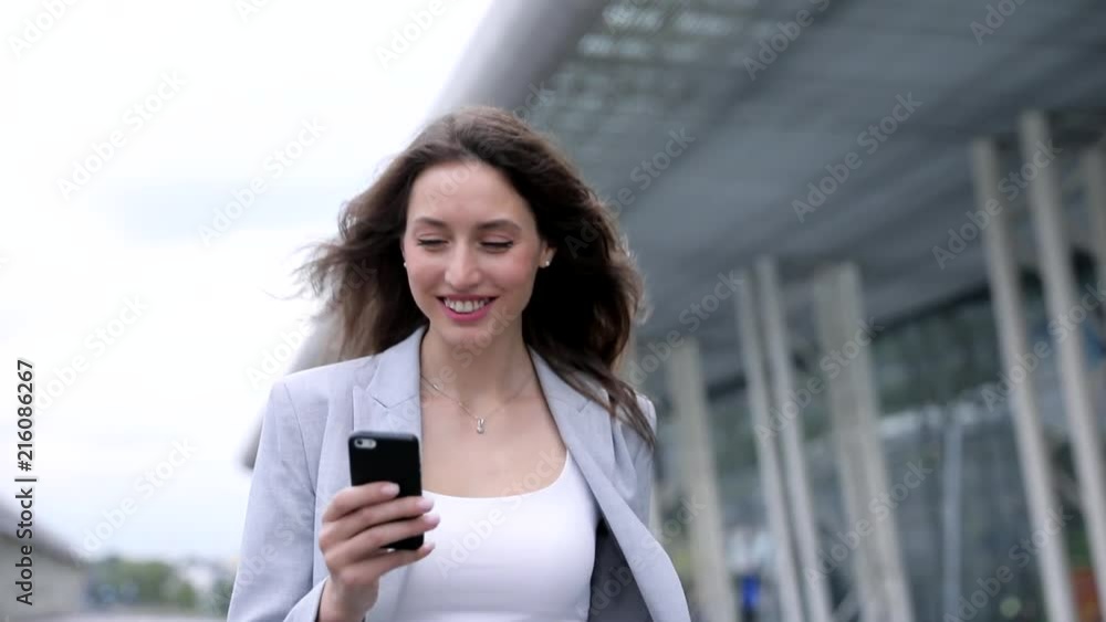 Portrait of Young Attractive Businesswoman Walking in the Town. Using her Modern Smartphone. Wearing Classical Suit. Huge Glass Airport in the Background. Businesswoman’s Charming Smile. Portrait of Young Attractive Businesswoman Walking in the Town. Using her Modern Smartphone. Wearing Classical Suit. Huge Glass Airport in the Background. Businesswoman’s Charming Smile.