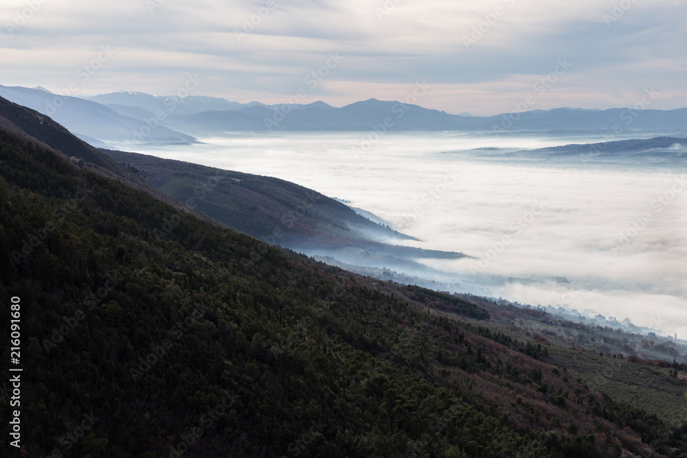 Beautiful aerial view of Umbria valley in a winter morning, with fog covering trees and houses , warm colors in the sky and layers of hills