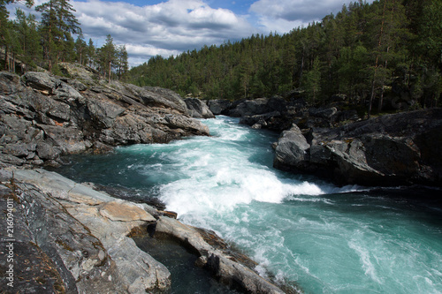 A stormy river stream with noise flows through the rocks and forest in Norway, clear transparent blue water