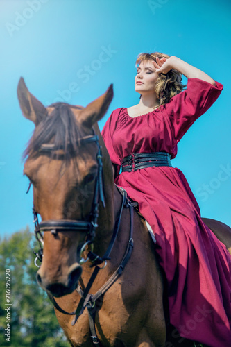 A young woman sits in a red dress is sitting on the back of a brown horse against a blue sky and clouds. Hand at the face.