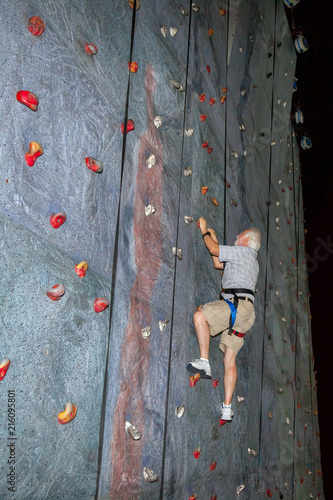 Elderly Man With White Hair and Beard Climbs a Rock Wall Simulation at Night at a Theme Park