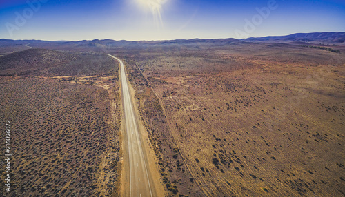 Rural road passing through dry land with scarce vegetation on bright sunny day - aerial panorama