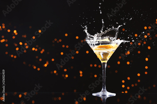 Glass with splashing cocktail and slice of orange against blurred lights