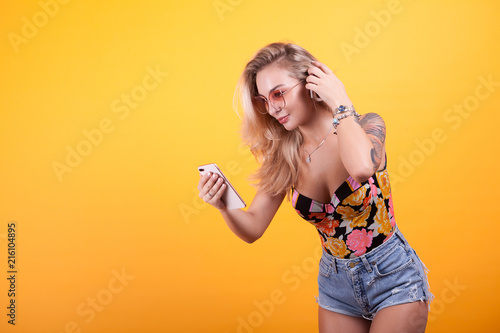 Smiling girl looking at phone screen in her hands with sunglasses in studio over yellow background