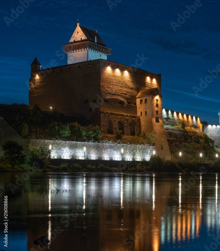Night view of Narva castle with the tower of High Herman, Narva, Estonia. The castle has a beautiful backlight. The castle is reflected in the water of the Narova River