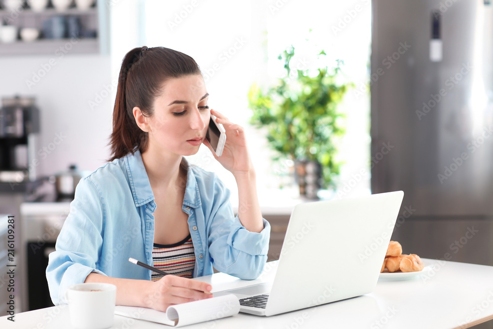 Young female freelancer talking on phone while working with laptop in kitchen Young female freelancer talking on phone while working with laptop in kitchen