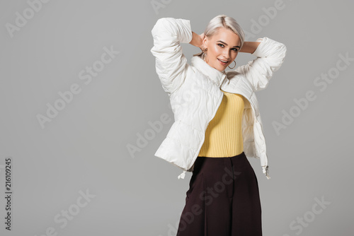 happy female model in stylish autumn jacket posing with raised arms isolated on grey background