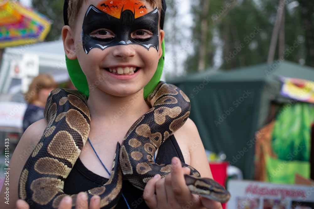 Child with the picture on the face of batman holding in the hands of the snake on ethno festival