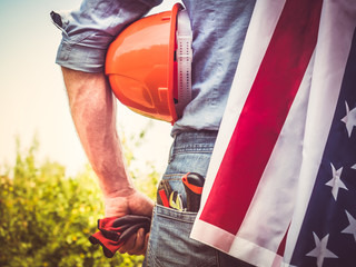 Attractive man in work clothes, holding tools and a US flag in his hands and looking into the distance against the background of trees, blue sky and sunset. View from the back. Labour Day Concept