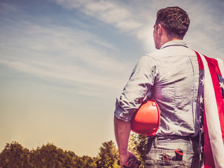 Attractive man in work clothes, holding tools and a US flag in his hands and looking into the distance against the background of trees, blue sky and sunset. View from the back. Labour Day Concept