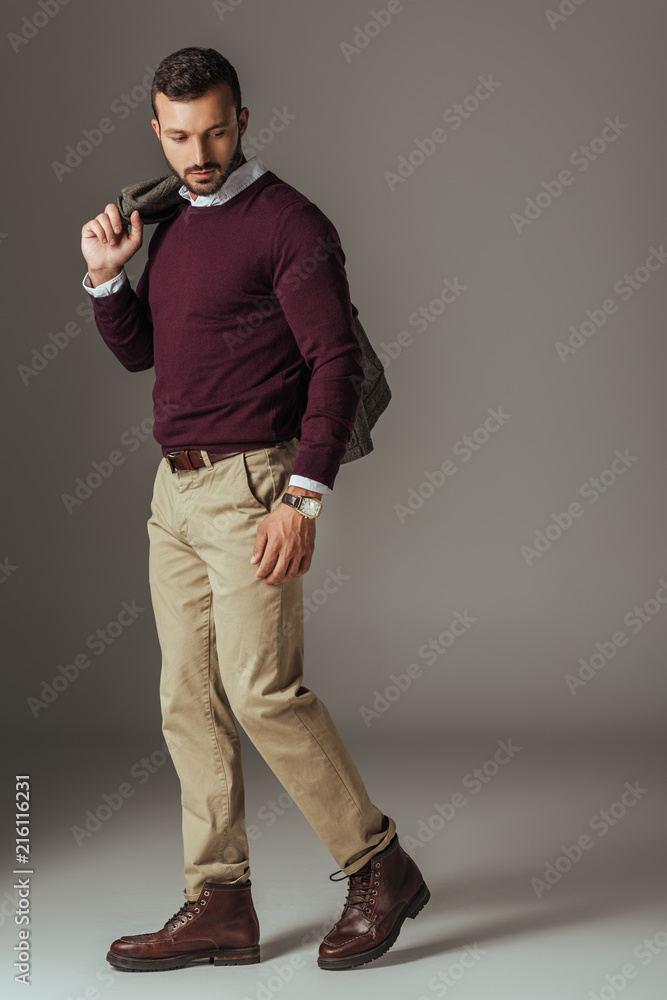 fashionable man posing in beige pants and burgundy sweater with autumn jacket on shoulder, on grey fashionable man posing in beige pants and burgundy sweater with autumn jacket on shoulder, on grey