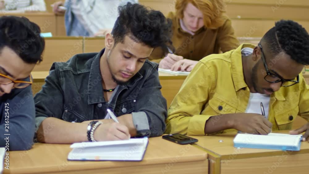 Tracking shot of three young men of different ethnicities sitting next to each other at desk and writing in notebooks during lecture at university Tracking shot of three young men of different ethnicities sitting next to each other at desk and writing in notebooks during lecture at university