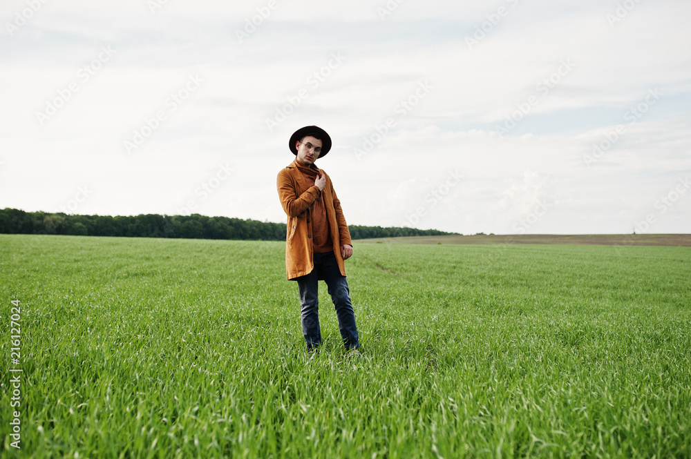 Stylish man in glasses, brown jacket and hat posed on green field. Stylish man in glasses, brown jacket and hat posed on green field.