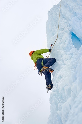 Alpinist man with ice tools axe in orange helmet climbing a lar