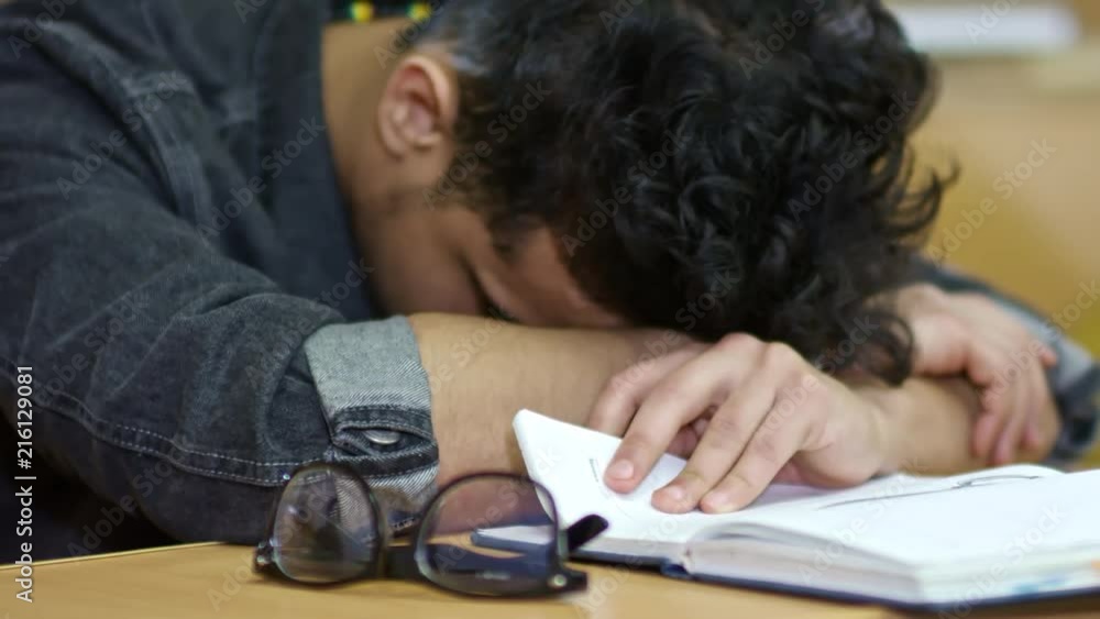 Close up shot of exhausted young student sleeping on notebook during lecture at university, his glasses lying near him on desk Close up shot of exhausted young student sleeping on notebook during lecture at university, his glasses lying near him on desk