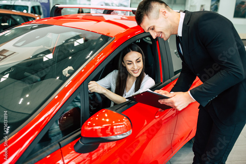 Salesman of dealership shows tablet with documents for familiarization to beautiful smiling woman, girl sitting inside a red car. © My Ocean studio