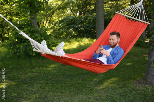 Young man with mobile phone resting in hammock outdoors