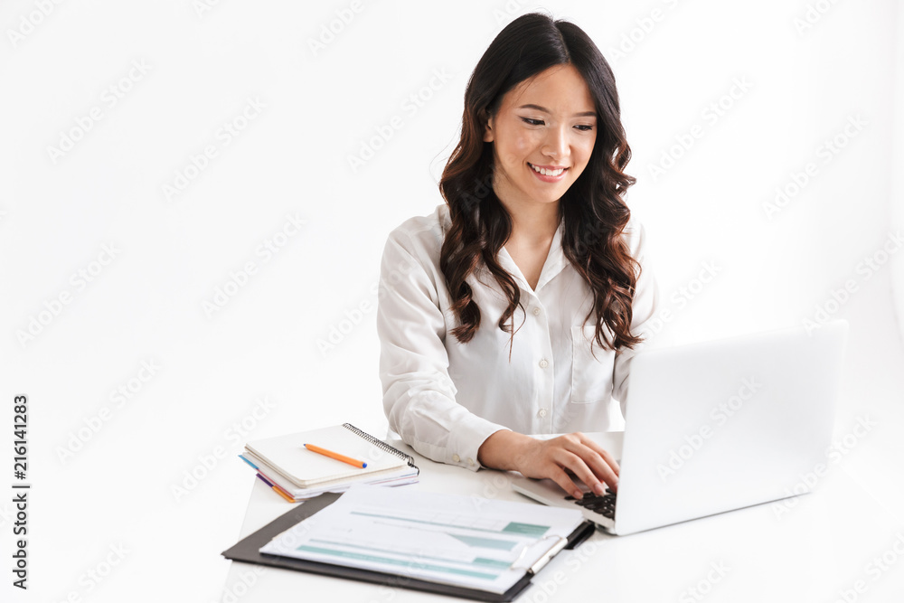 Photo of successful asian woman with long dark hair sitting at table and working with documents and laptop, isolated over white background in studio Photo of successful asian woman with long dark hair sitting at table and working with documents and laptop, isolated over white background in studio