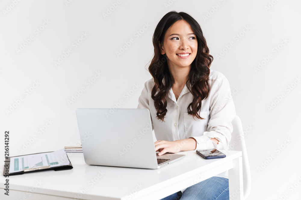 Photo of charming chinese businesswoman with long dark hair sitting at table and working with documents and laptop, isolated over white background in studio Photo of charming chinese businesswoman with long dark hair sitting at table and working with documents and laptop, isolated over white background in studio