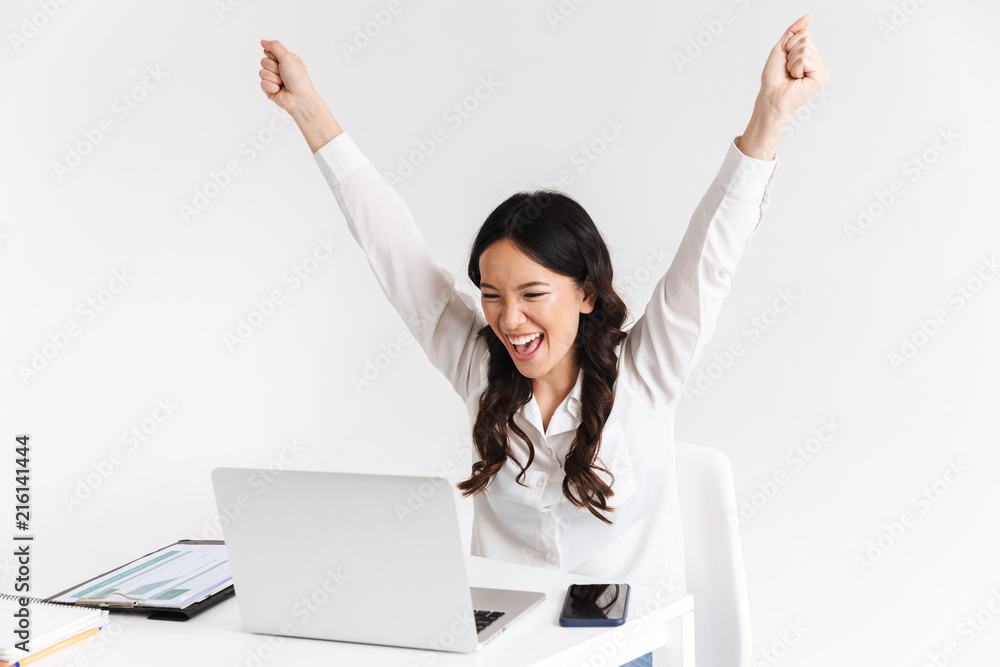 Photo of excited chinese businesswoman with long dark hair screaming with raised arms while working with documents and laptop, isolated over white background in studio Photo of excited chinese businesswoman with long dark hair screaming with raised arms while working with documents and laptop, isolated over white background in studio