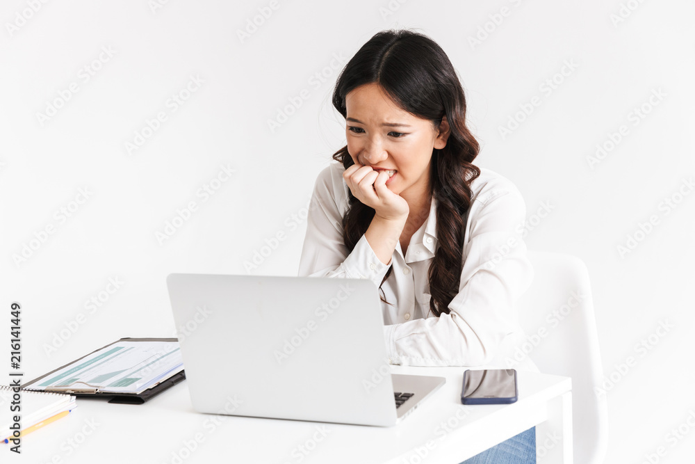 Photo of disappointed young asian business woman 20s wearing office clothing sitting at table with laptop and biting fist, isolated over white background Photo of disappointed young asian business woman 20s wearing office clothing sitting at table with laptop and biting fist, isolated over white background