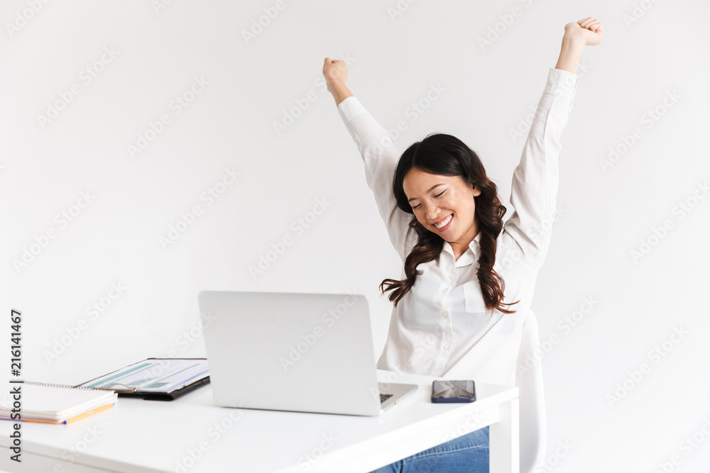 Photo of successful chinese businesslike woman with long dark hair smiling with raised arms while working with documents and laptop, isolated over white background Photo of successful chinese businesslike woman with long dark hair smiling with raised arms while working with documents and laptop, isolated over white background