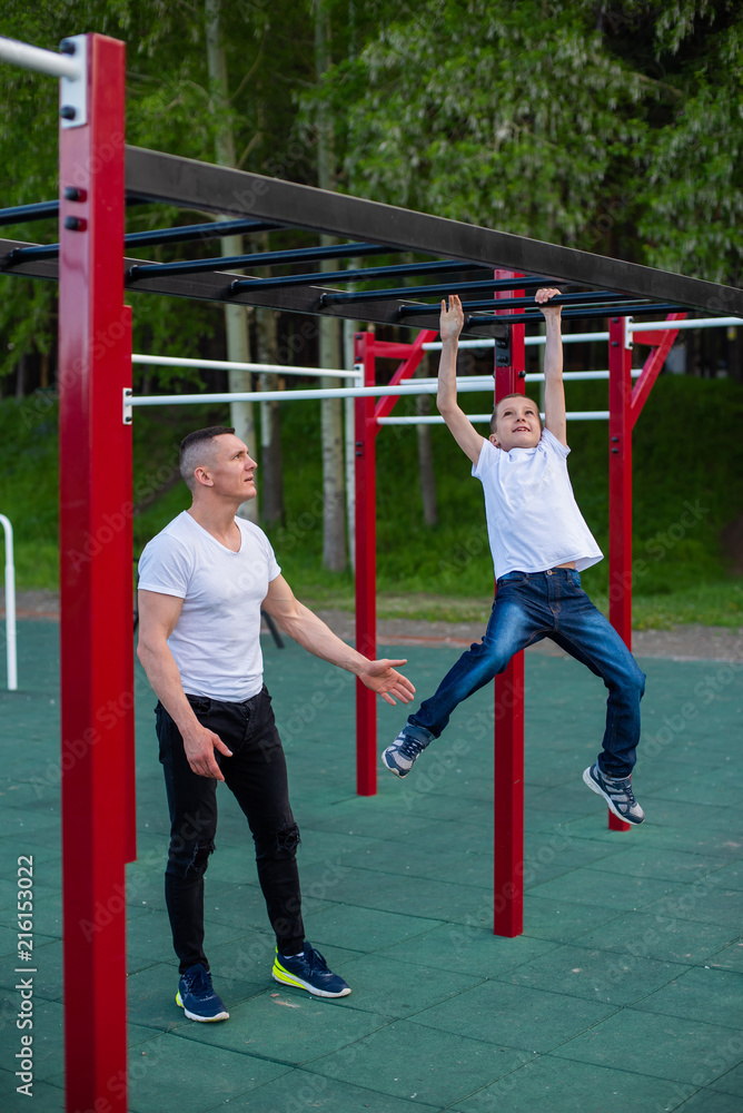 A strong athletic father insures the boy on sports equipment and teaches him to pull himself up on the horizontal bar in the summer park. Strong athletic father teaches his son to catch up on the bar.
